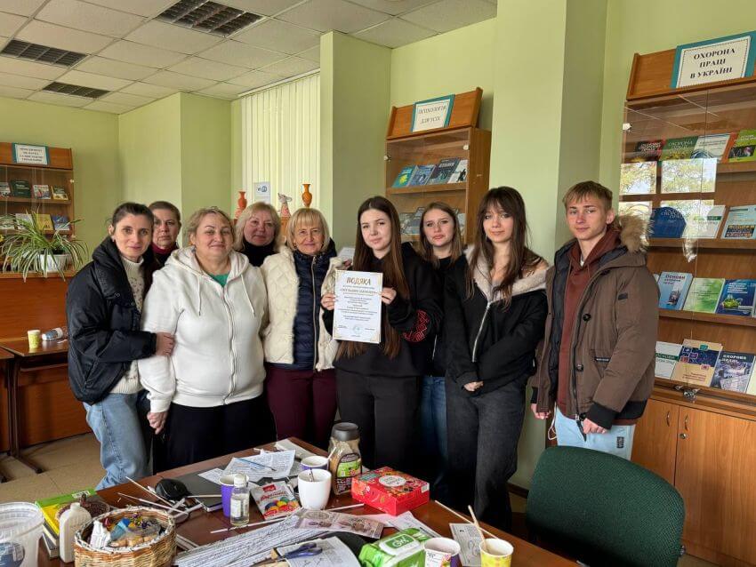 Групове фото учасників майстер-класу в бібліотеці. Студенти та організатори стоять разом, студентка коледжу Ульяна Чабанна тримає в руках подяку за участь у виставці «Світ наших захоплень». Поруч видно книжкові стелажі та стіл з матеріалами для творчості