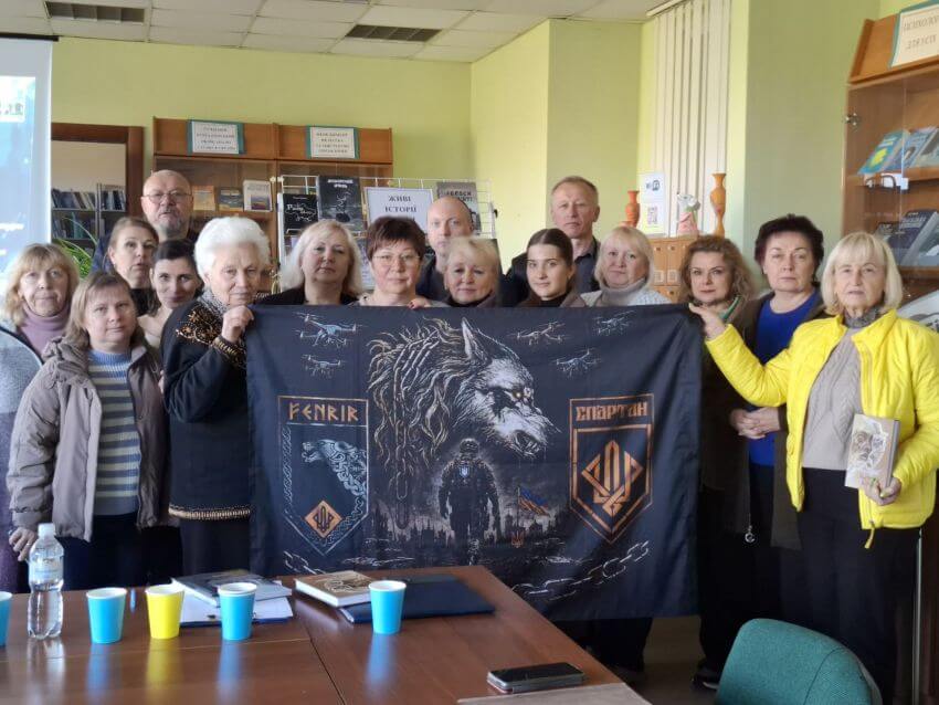 Учасники та гості творчої зустрічі в Науковій бібліотеці ДПУ з Ольгою Воробйовою тримають пам'ятний військовий прапор із символікою 3-ї бригади оперативного призначення Національної гвардії України «Спартан» (імені полковника Петра Болбочана), а також окремого підрозділу/групи «FENRIR» (з відповідним шевроном та зображенням вовка)