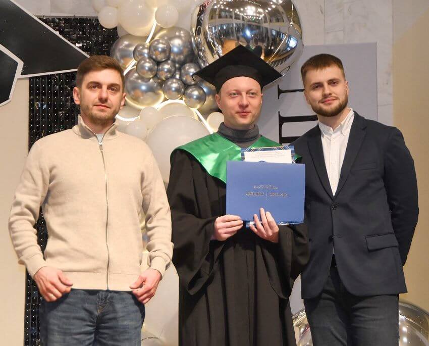 Випуск магістрів ДПУ. Троє молодих чоловіків стоять разом на фоні святкових декорацій із кульок. Центральний чоловік, Дмитро Бузинник, одягнений у мантію випускника та конфедератку, він тримає в руках синю папку з дипломом